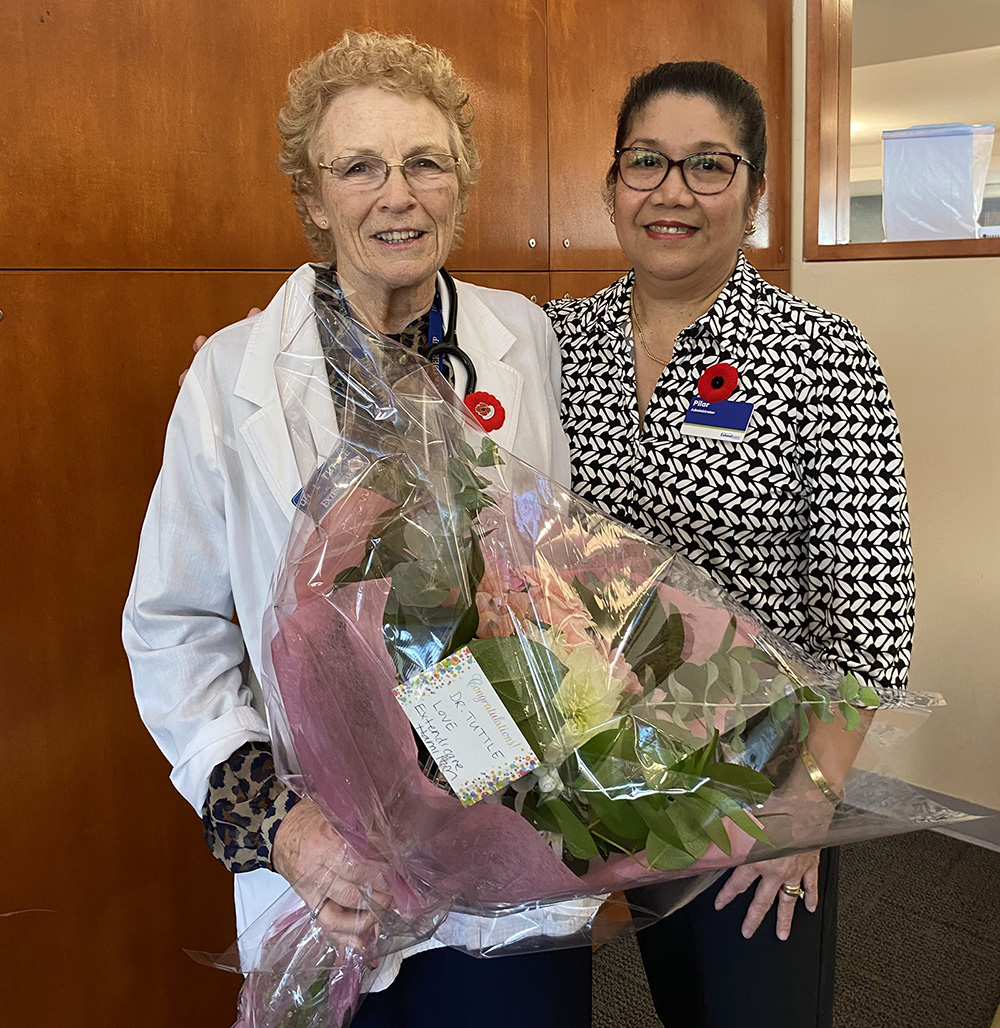 Two women stand side by side indoors, both smiling. One holds a large bouquet wrapped in pink paper, and both wear poppy pins on their clothing.