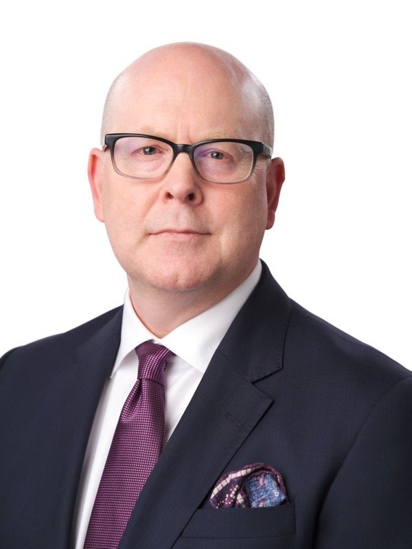 Bald man wearing glasses, a dark suit, white shirt, and purple tie, standing against a plain white background.