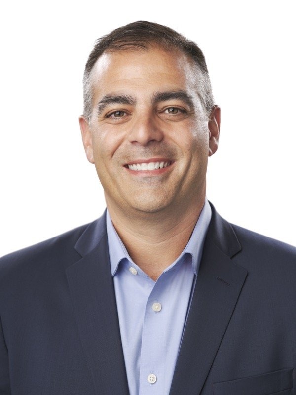 A man with short dark hair wearing a navy blazer and light blue shirt, smiling at the camera against a plain white background.