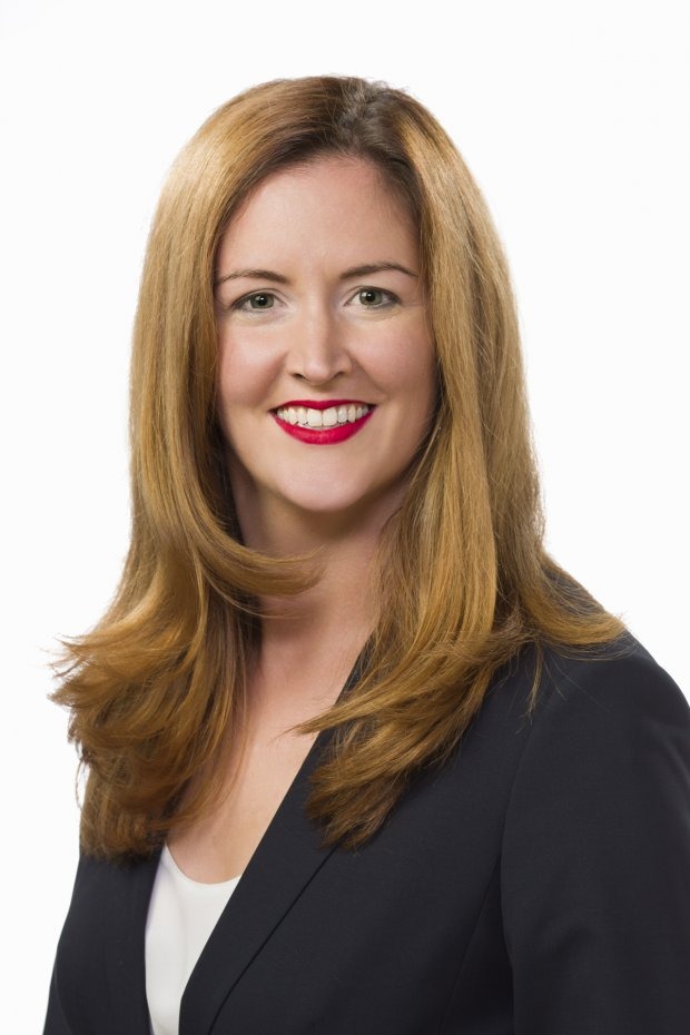 Woman with long auburn hair wearing a dark blazer and white top, smiling at the camera against a plain white background.