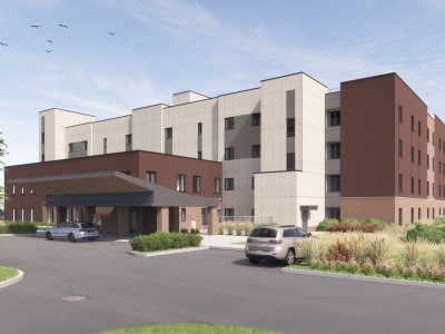 A four-story modern building with light and dark brown exterior walls, an entrance canopy, parked cars, and landscaped grounds under a partly cloudy sky.