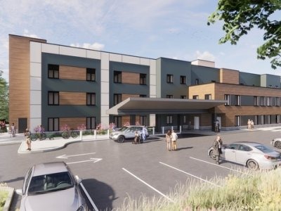 A modern three-story building with large windows, a covered entrance, parked cars, and people walking in the parking lot under a partly cloudy sky.