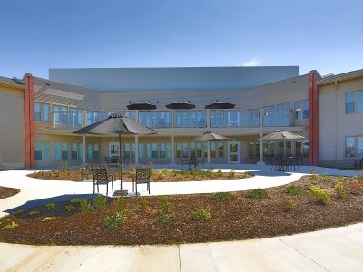 A modern two-story building with large windows and an outdoor courtyard featuring tables, chairs, umbrellas, and landscaped garden beds.