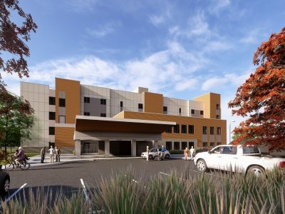 A modern, multi-story building with a covered entrance, cars parked in front, and people walking nearby under a partly cloudy sky.