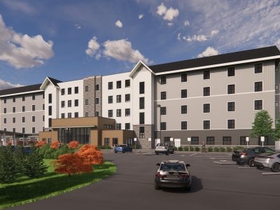 A four-story modern apartment building with parked cars in front and landscaping including green grass, trees, and shrubs under a partly cloudy sky.