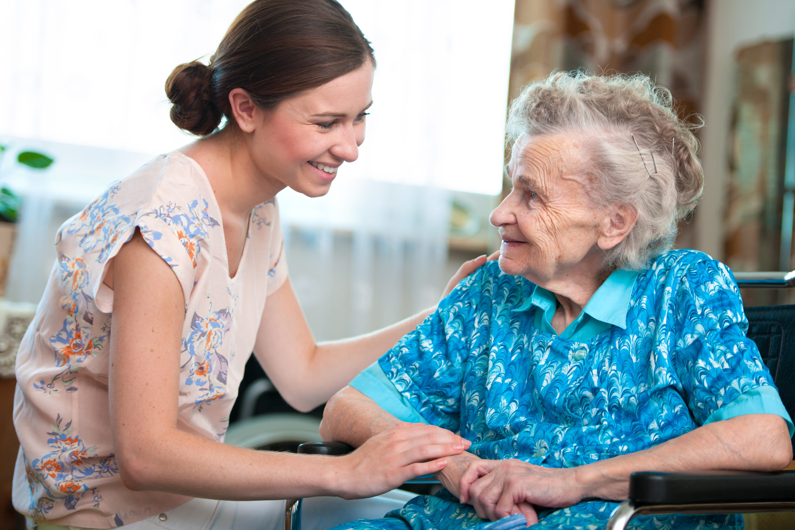 A young woman smiles and holds hands with an elderly woman sitting in a wheelchair indoors.