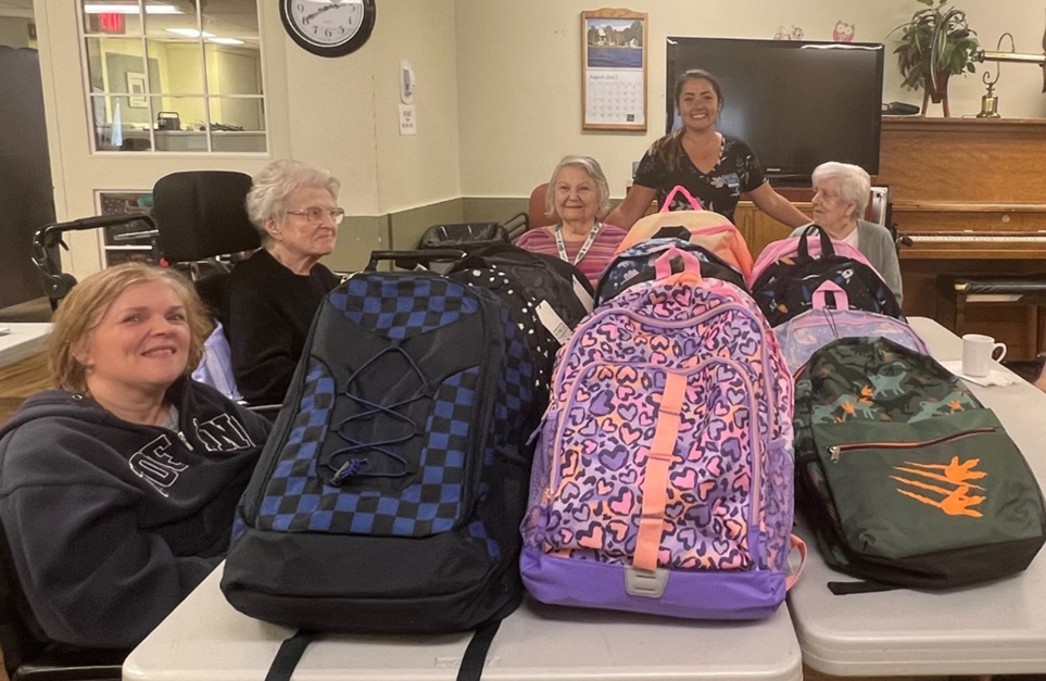 Five women sit and stand around a table with several colorful backpacks. The setting appears to be a community room with a clock, calendar, and piano in the background.