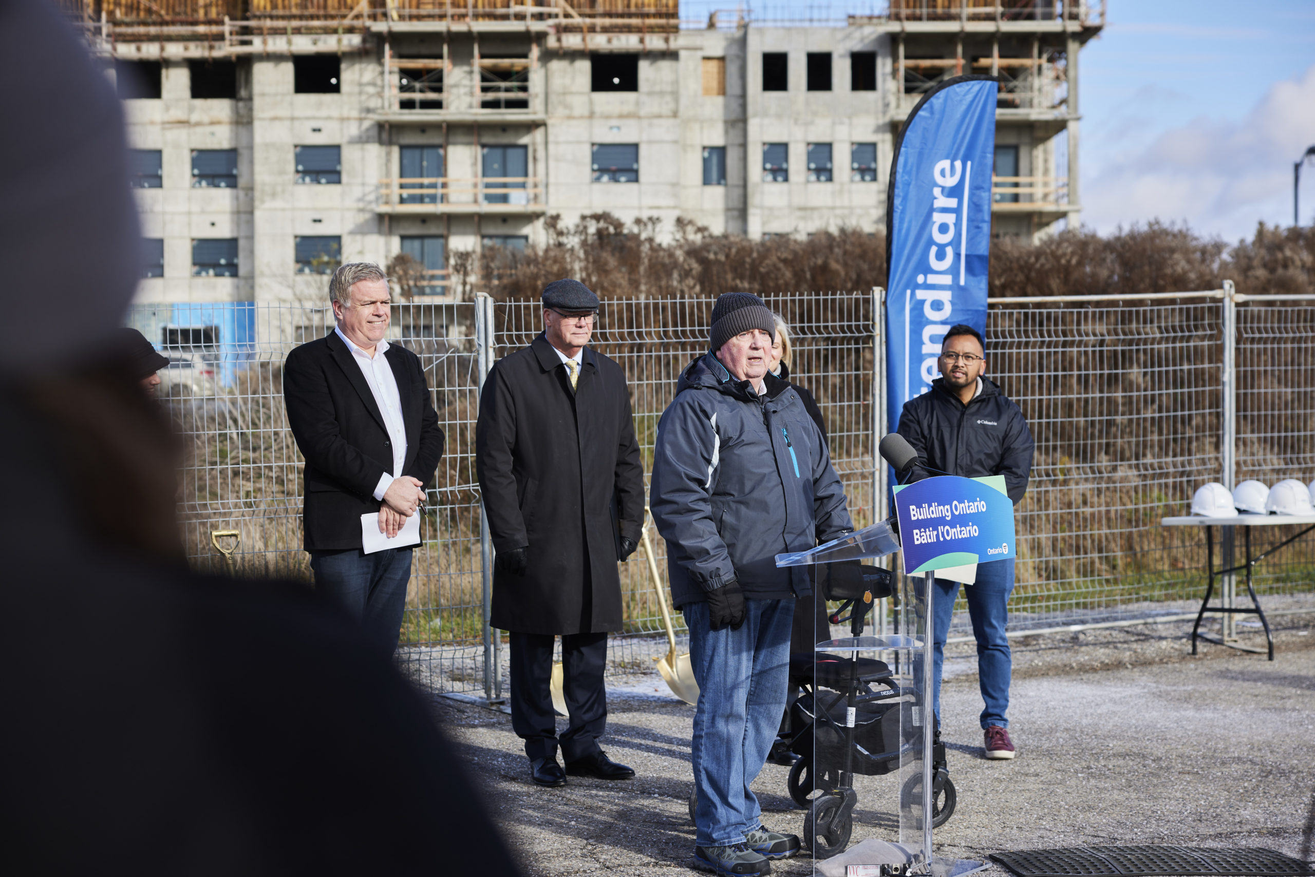 A speaker addresses an outdoor press event at a construction site, with three people standing behind and construction in the background.