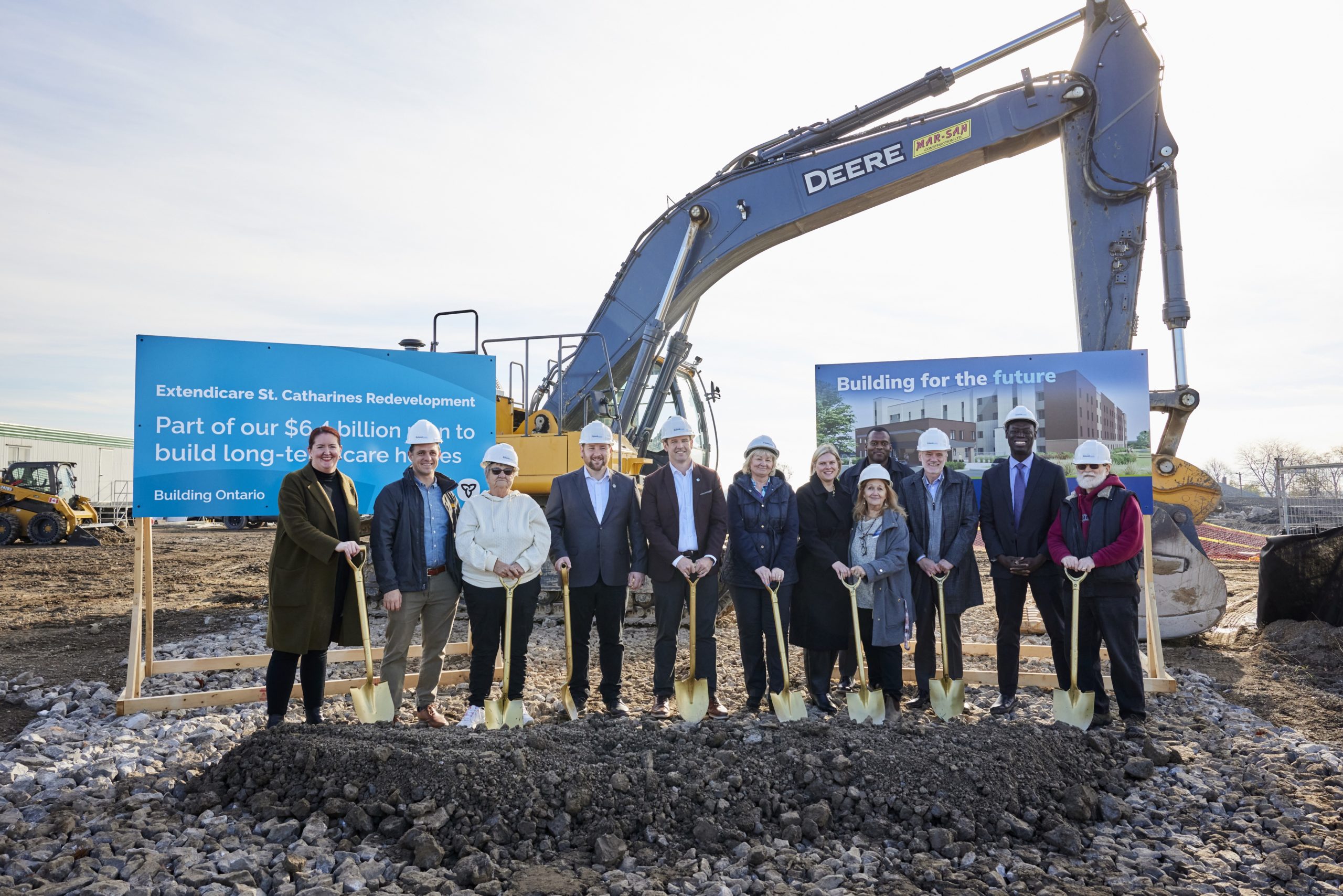 A group of people in hard hats stand with shovels at a groundbreaking ceremony in front of construction equipment and project signs.