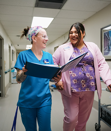 Two healthcare workers in scrubs walk down a hallway, smiling and looking at a large open binder together.