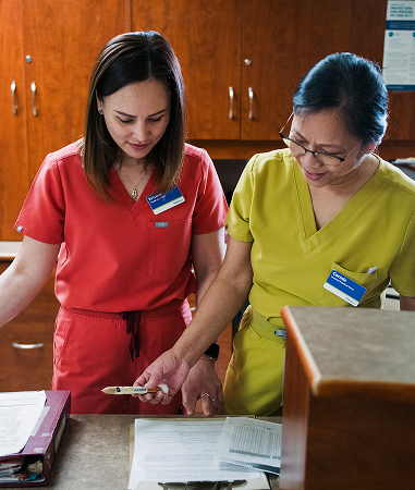 Two healthcare workers in scrubs review documents together at a counter in a medical office setting.