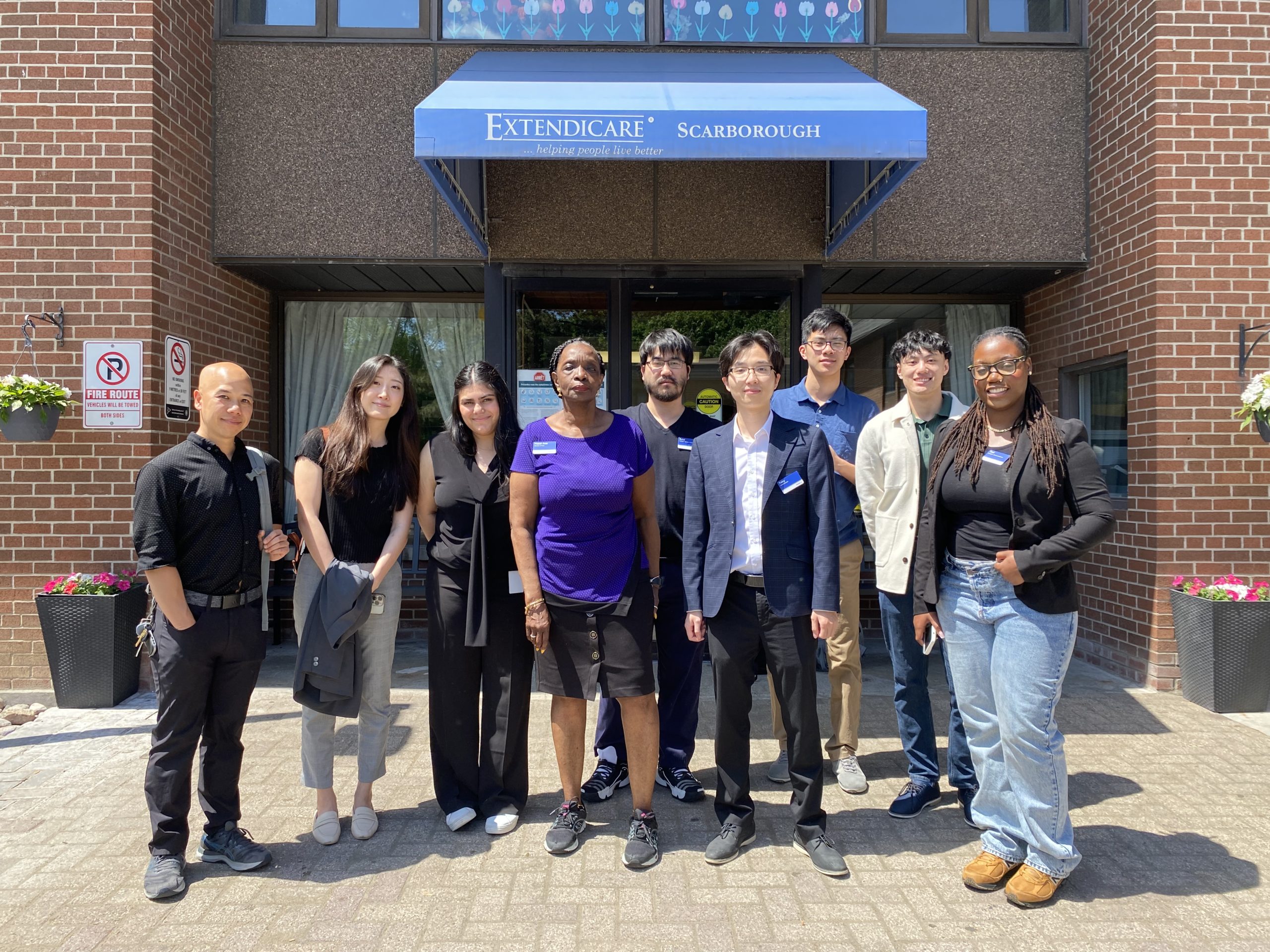 A group of nine people stands in front of the Extendicare Scarborough entrance on a sunny day, posing for a photo.