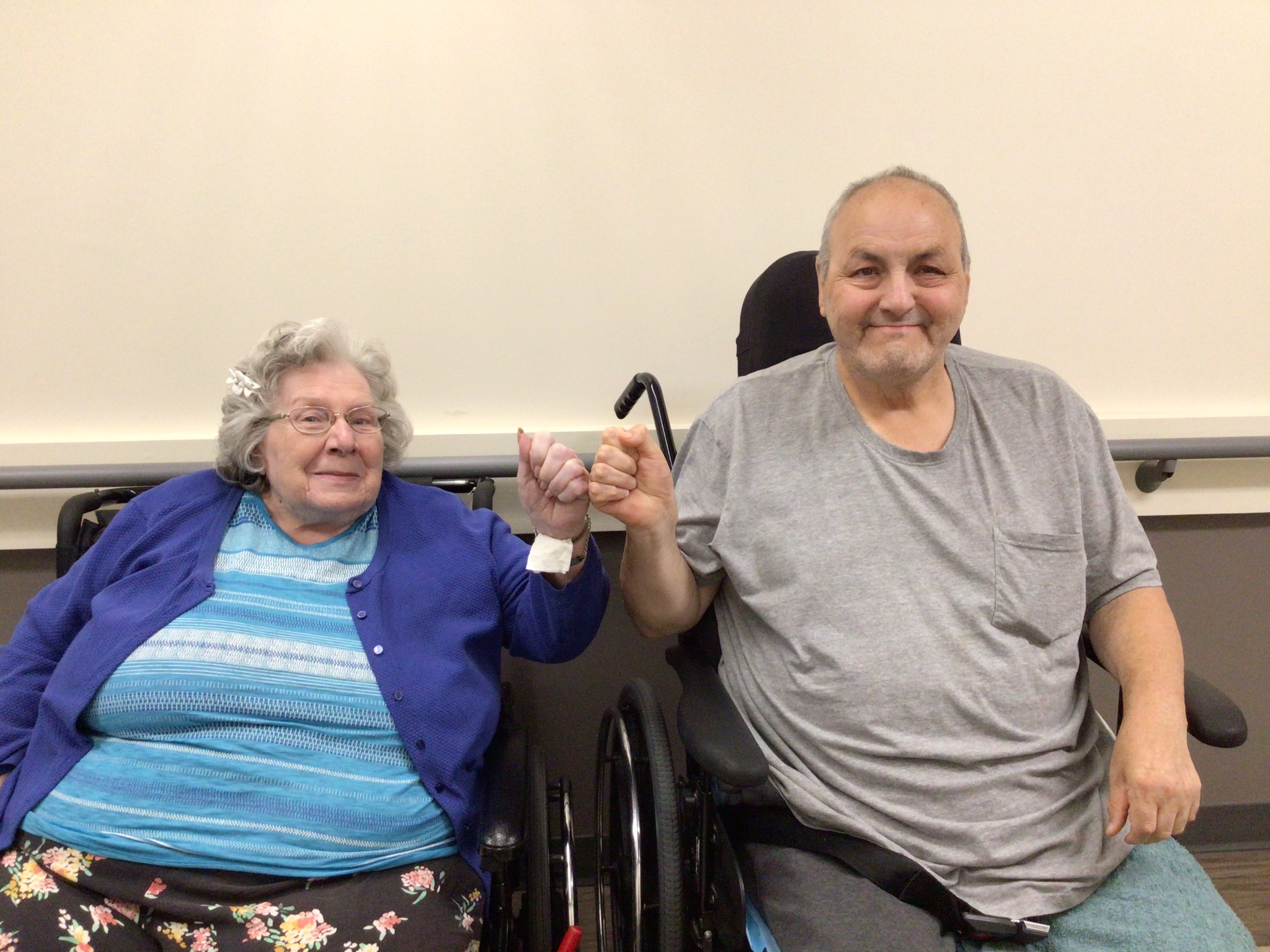 An elderly woman and man, both in wheelchairs, sit side by side holding hands and smiling at the camera in front of a plain background.