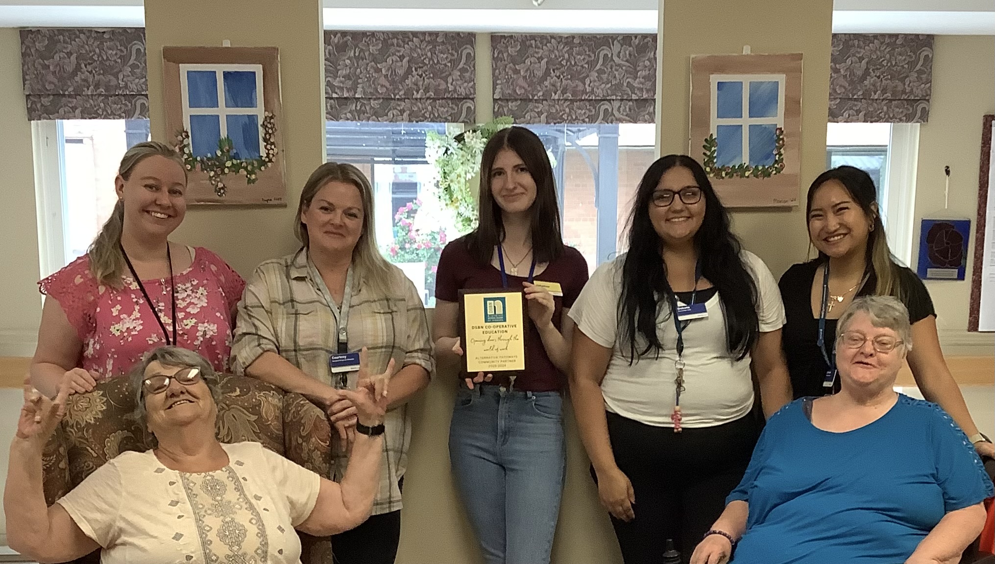 Six women, including two seated older women and four standing younger women, pose indoors. The woman in the center holds a recognition plaque. All are smiling.