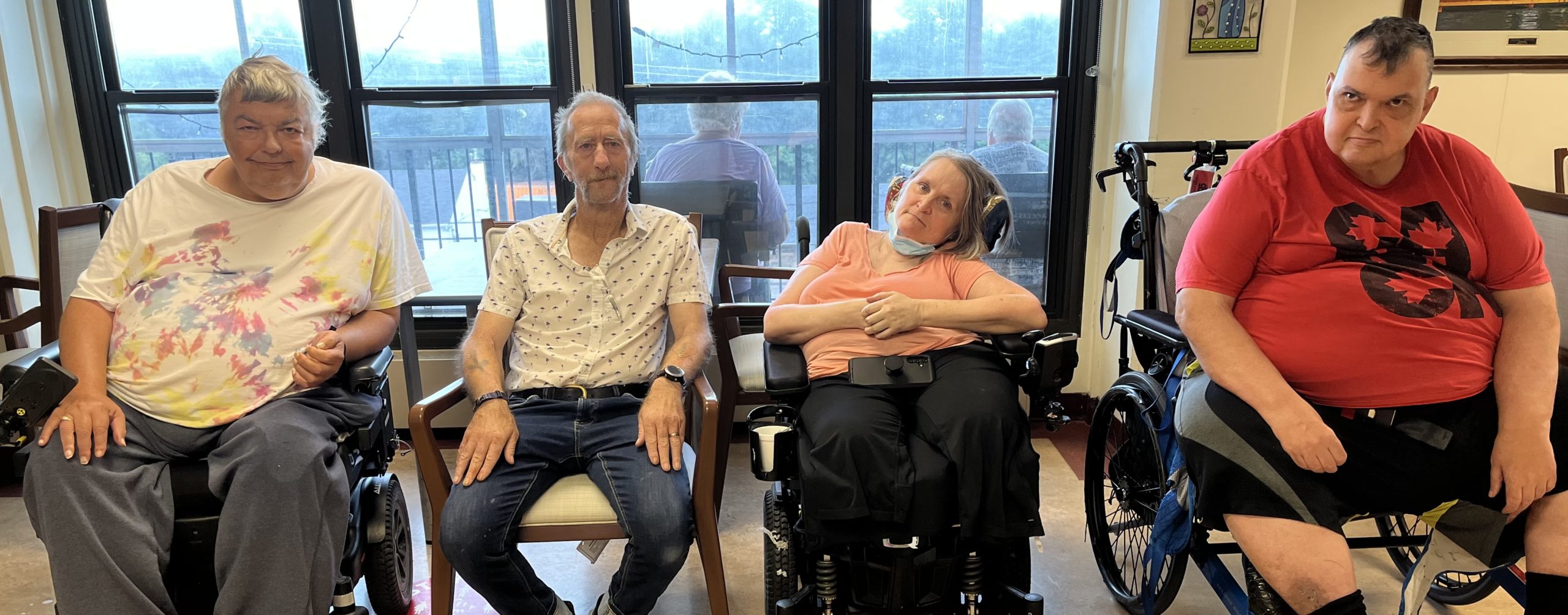 Four adults, three in wheelchairs and one in a chair, sit indoors in front of large windows, looking toward the camera.
