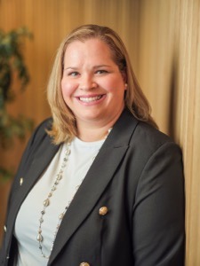 A woman with blonde hair wearing a dark blazer, light top, and necklace, smiling while standing in front of a wooden wall with a plant in the background.