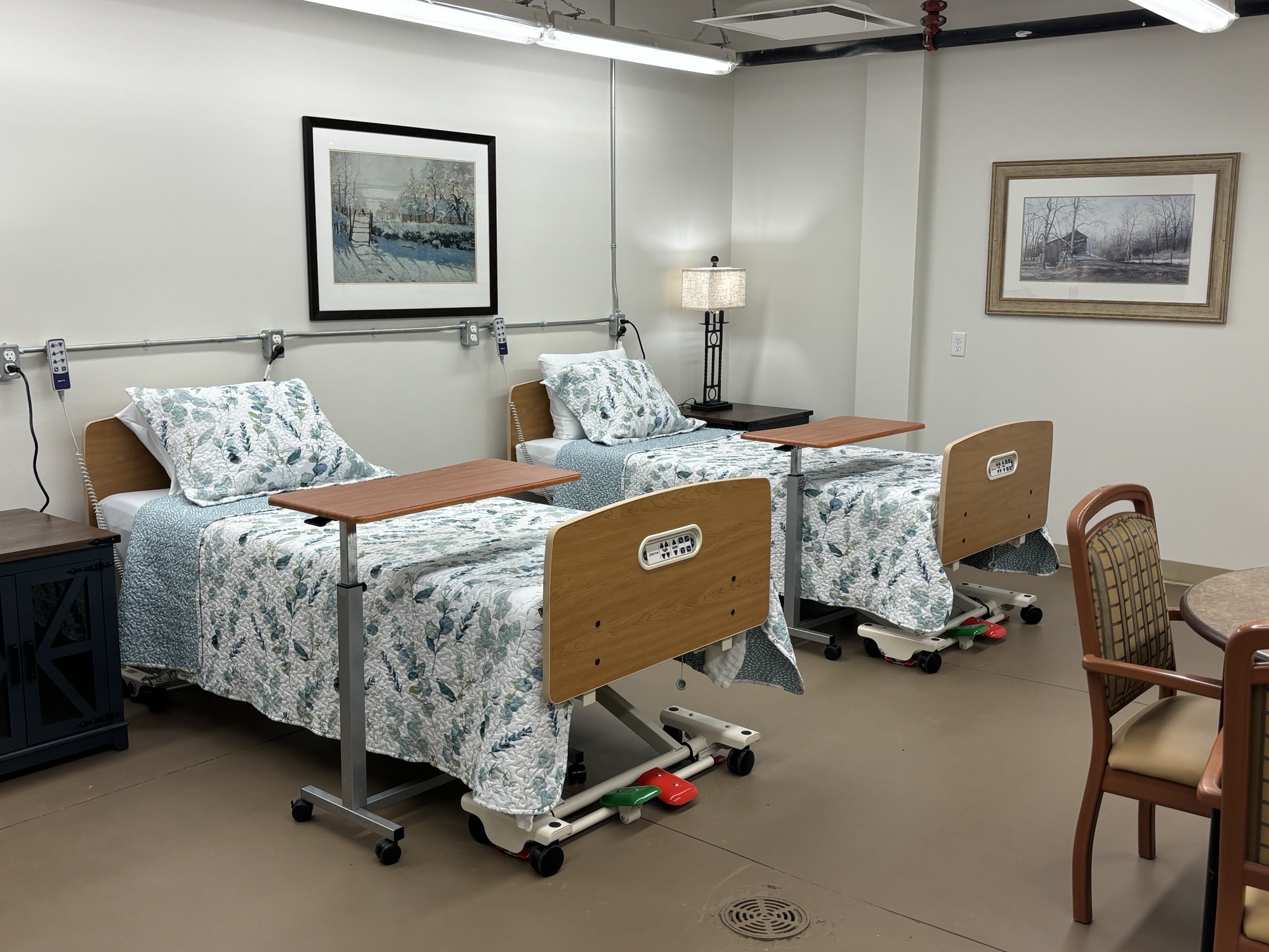 Two hospital beds with floral bedding are side by side in a clean, well-lit room, each with an overbed table. The room has framed wall art, a lamp, and a small table with chairs.