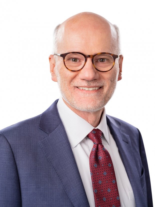 A man with glasses, a beard, and short hair is wearing a blue suit, white shirt, and red patterned tie, smiling against a white background.