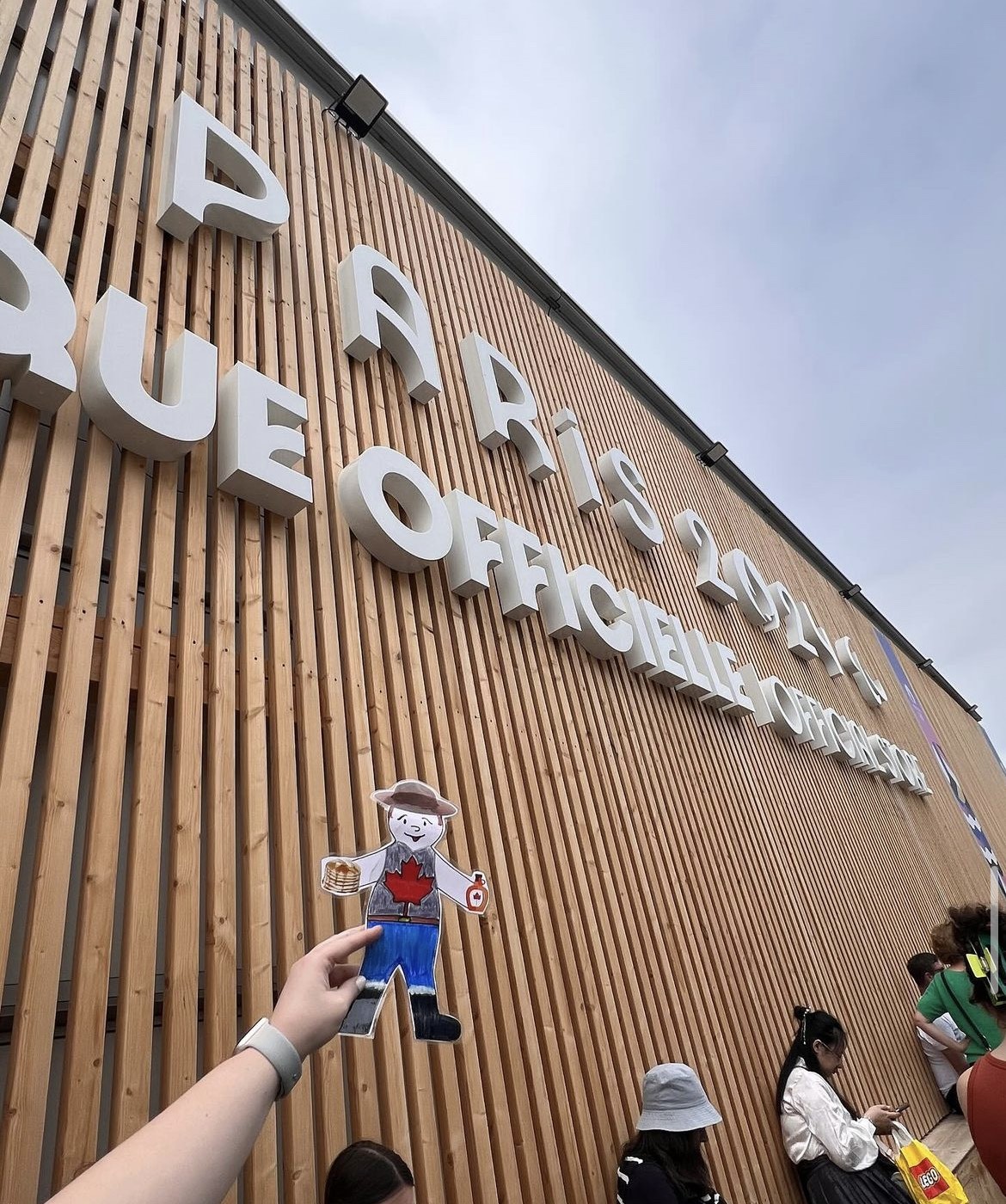 A hand holds a paper cutout of a character in front of a wooden building with large white letters reading “PARIS 2024 - PARQUE OFFICIELLE - OFFICIAL PARK.” Several people stand below.