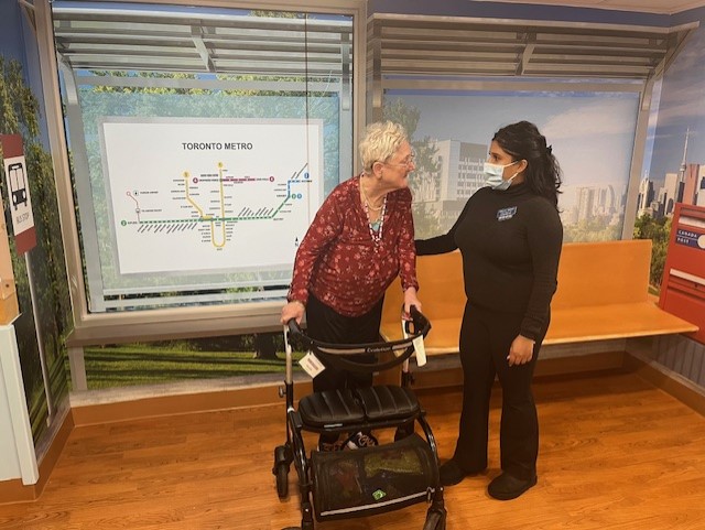 An elderly woman with a walker stands beside a caregiver in front of a Toronto Metro map and a bench in a brightly lit room.