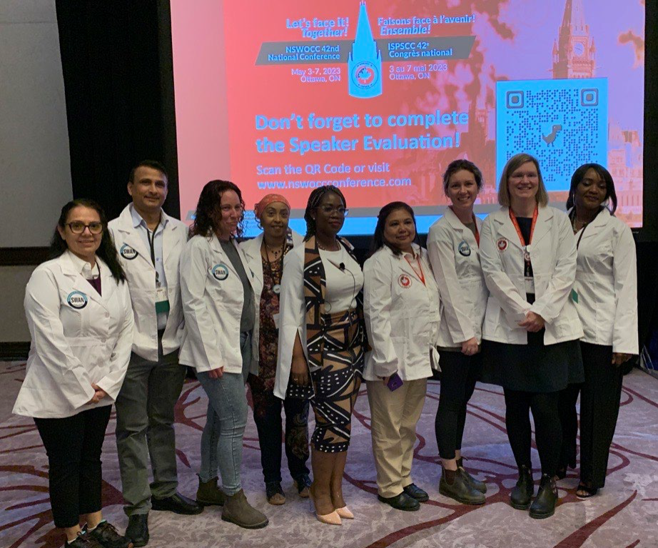 A group of ten people, most wearing white lab coats, pose together in front of a screen displaying information about a conference and a QR code.
