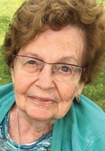 Elderly woman with short curly hair and glasses, wearing a blue top and cardigan, looking at the camera outdoors.