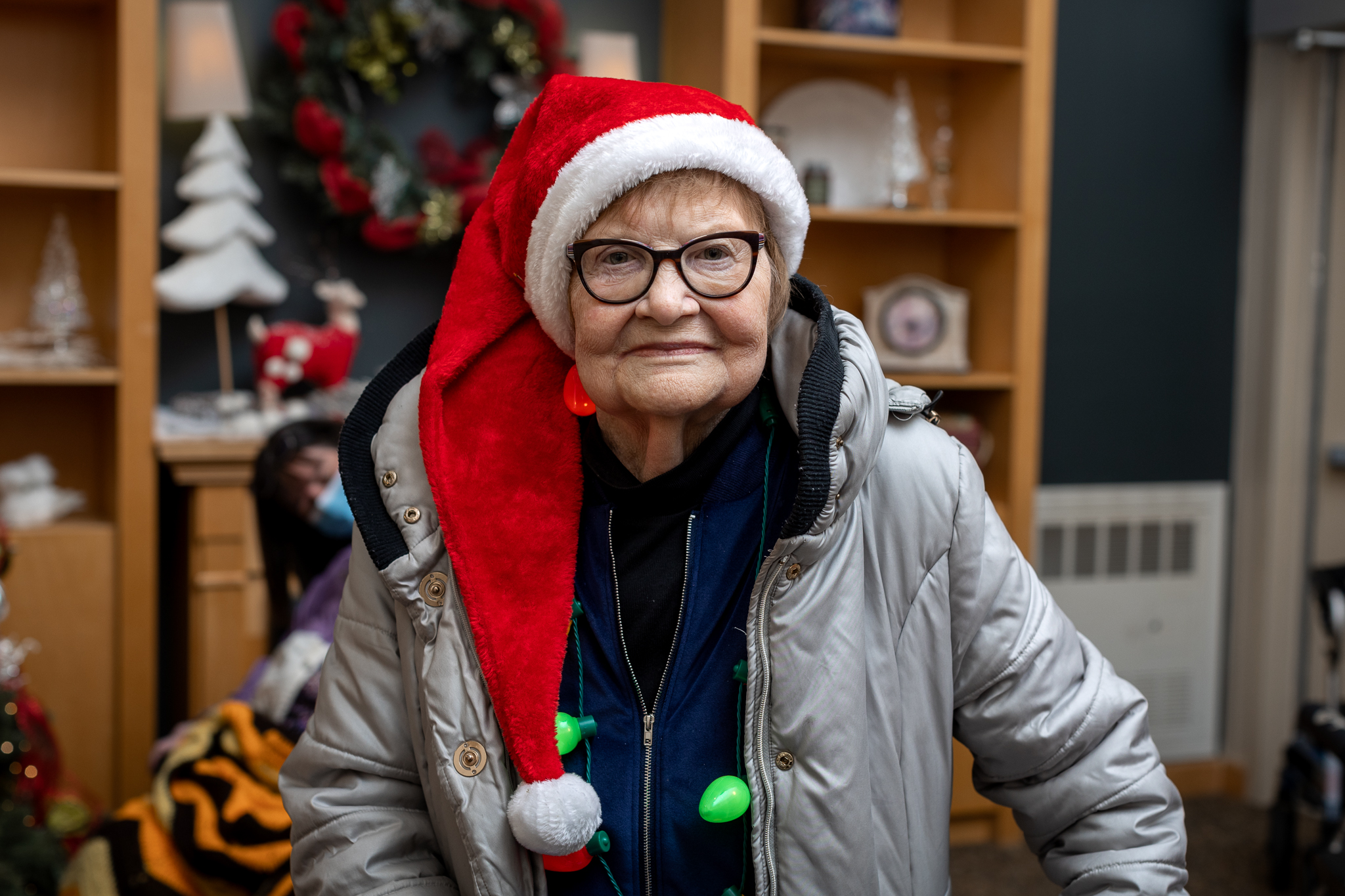 An elderly person wearing glasses, a silver coat, and a red Santa hat stands indoors with holiday decorations in the background.