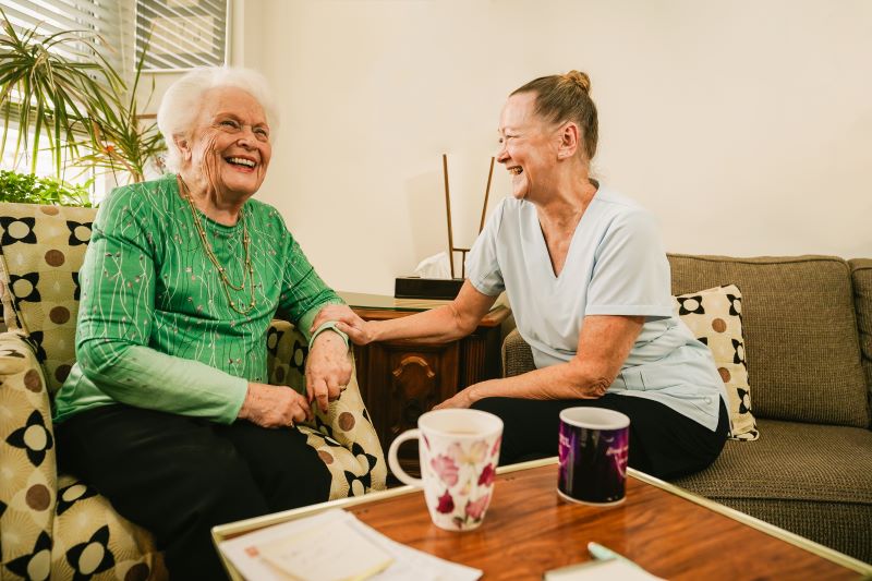 Two women sit on a couch smiling and holding hands, with coffee mugs and papers on the table in front of them in a brightly lit living room.