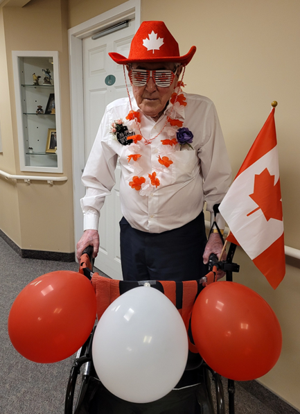 Elderly man wearing a red cowboy hat, Canadian flag sunglasses, and floral lei, standing behind a walker decorated with red and white balloons and a Canadian flag.