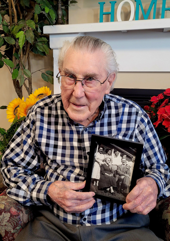 Elderly man in a plaid shirt sits indoors, holding a black-and-white family photo in his lap. Bright flowers and a "HOME" sign are visible in the background.