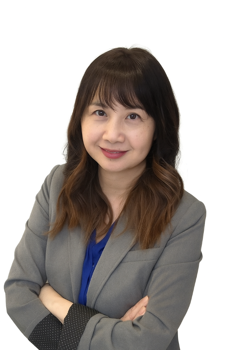 A woman with long brown hair wearing a gray blazer and blue blouse stands with arms crossed, smiling at the camera against a white background.