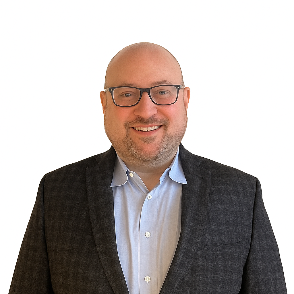 A bald man with glasses, wearing a dark checkered blazer and light blue shirt, stands smiling against a plain white background.