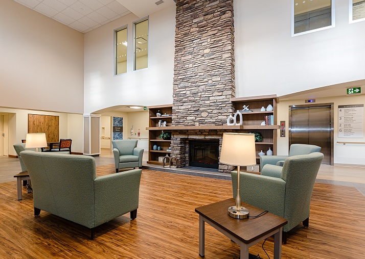 A spacious lobby with green armchairs, wooden tables, and a large stone fireplace; elevator and hallway visible in the background.