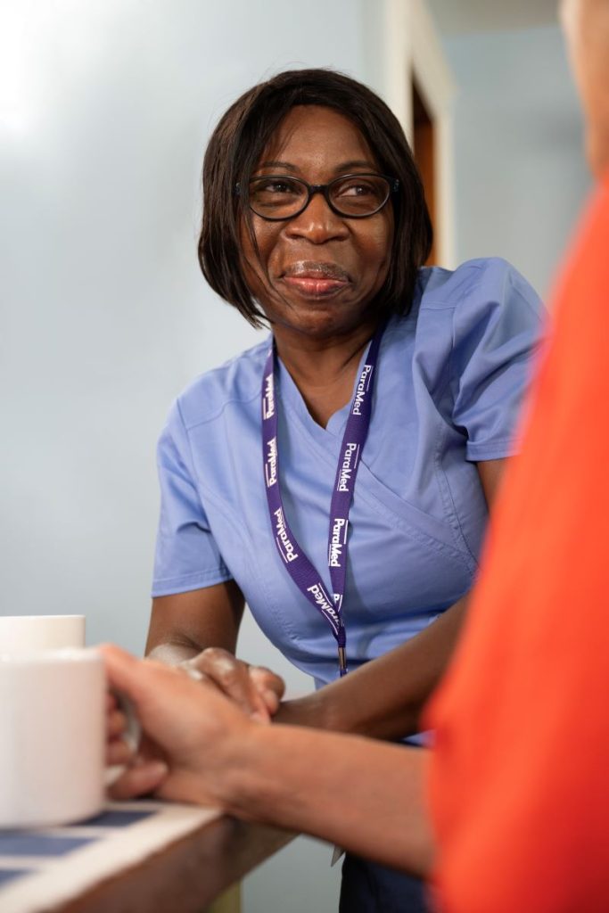 A woman in blue scrubs and a lanyard smiles while sitting at a table with another person, both holding mugs.
