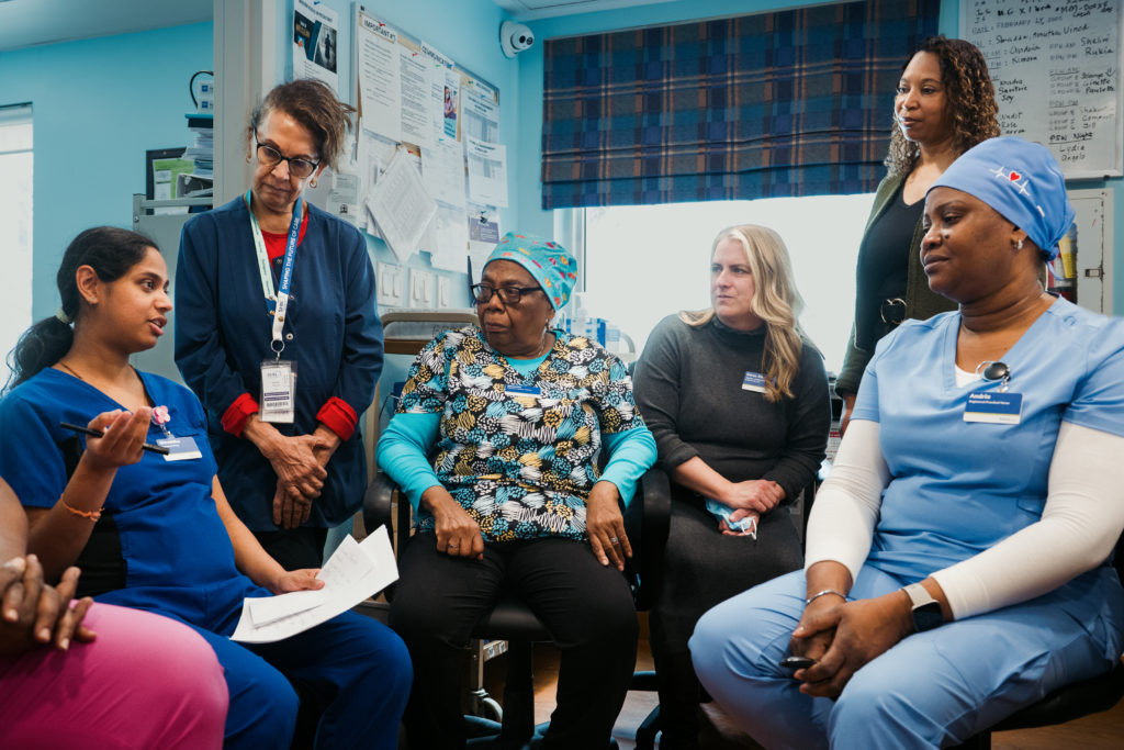 A group of healthcare workers in scrubs and uniforms sit and stand together in a medical office, engaged in discussion.