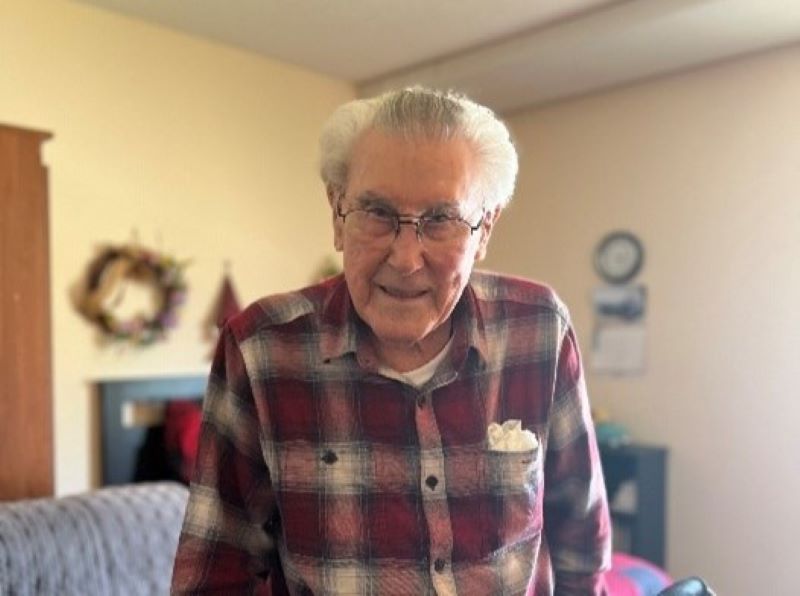 An elderly man with gray hair and glasses wears a red plaid shirt and smiles while standing in a cozy room with simple decor in the background.