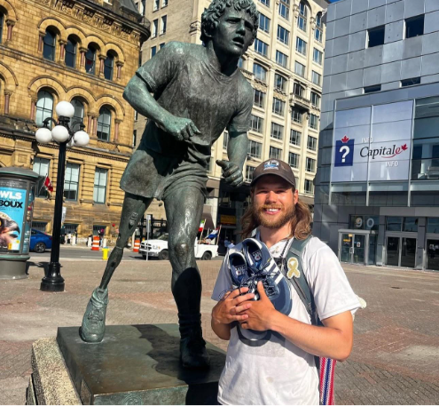 A man stands in front of a running statue in an urban square, holding a pair of sneakers and smiling at the camera.