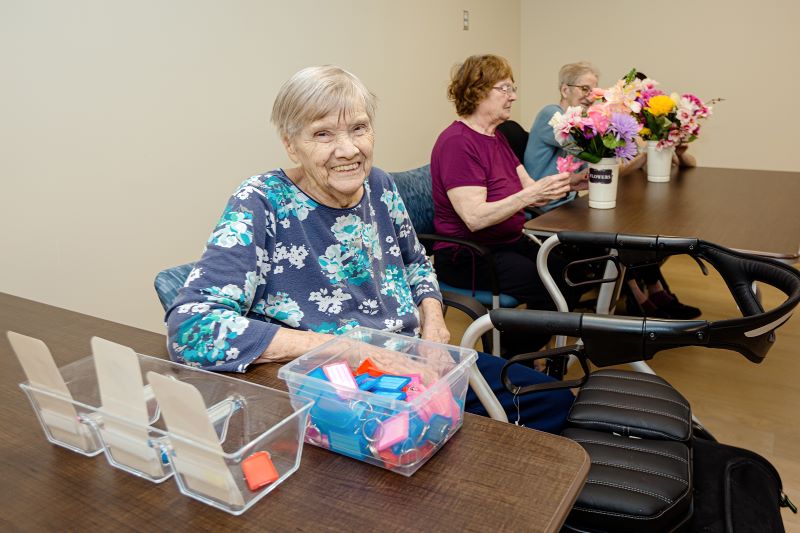 Three elderly women sit at a table; one smiles at the camera with sorting materials, while two others work with flower arrangements in the background.