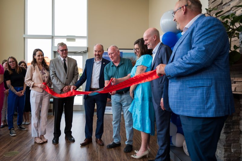 A group of people stand in a semi-circle as a man cuts a red ribbon with large scissors at an indoor event.